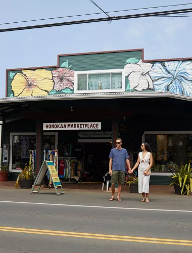 Couple holding hands outside the Honokaa Marketplace, a colorful building decorated with hibiscus murals, on a sunny day.