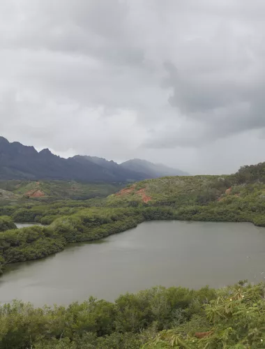 A serene fishpond surrounded by lush green mountains under a cloudy sky.