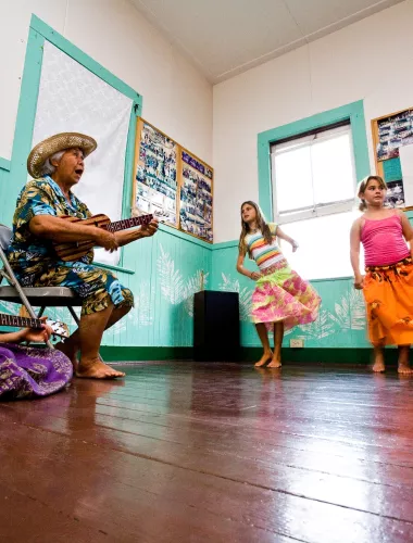 Children learning to hula as older woman plays ukelele.