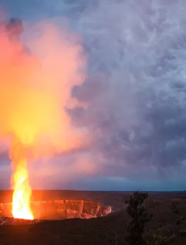 Glowing lava erupts from a volcanic crater, lighting up the dark sky with vibrant orange hues against dramatic storm clouds.