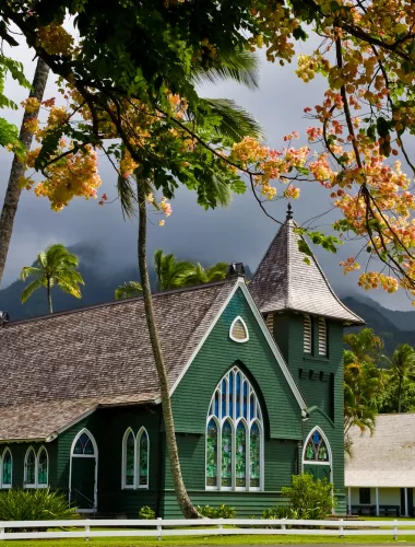 Waioli Mission House and Church nestled among palm trees with a green mountain backdrop.