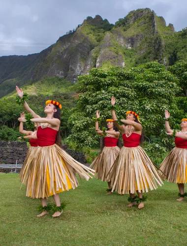 Traditional hula dancers perform on green grass with a backdrop of tropical trees and dramatic mountain cliffs.