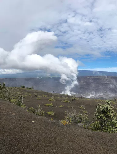 A landscape of Hawai‘i Volcanoes National Park featuring a steaming volcanic crater surrounded by barren terrain and sparse greenery under a cloudy sky.