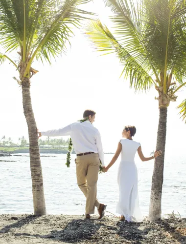 Couple holding hands under palm trees by the ocean, gazing into the distance on a sunny day.