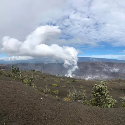 A landscape of Hawai‘i Volcanoes National Park featuring a steaming volcanic crater surrounded by barren terrain and sparse greenery under a cloudy sky.