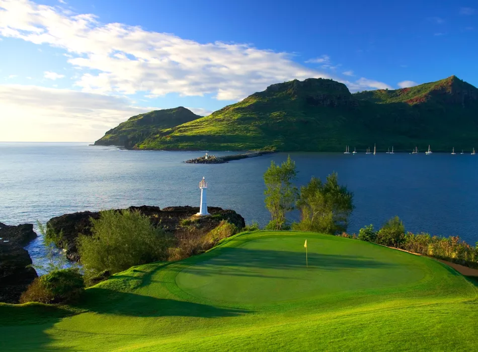 Golf course with a lush green putting green overlooking the ocean and sailboats near a mountainous coastline.