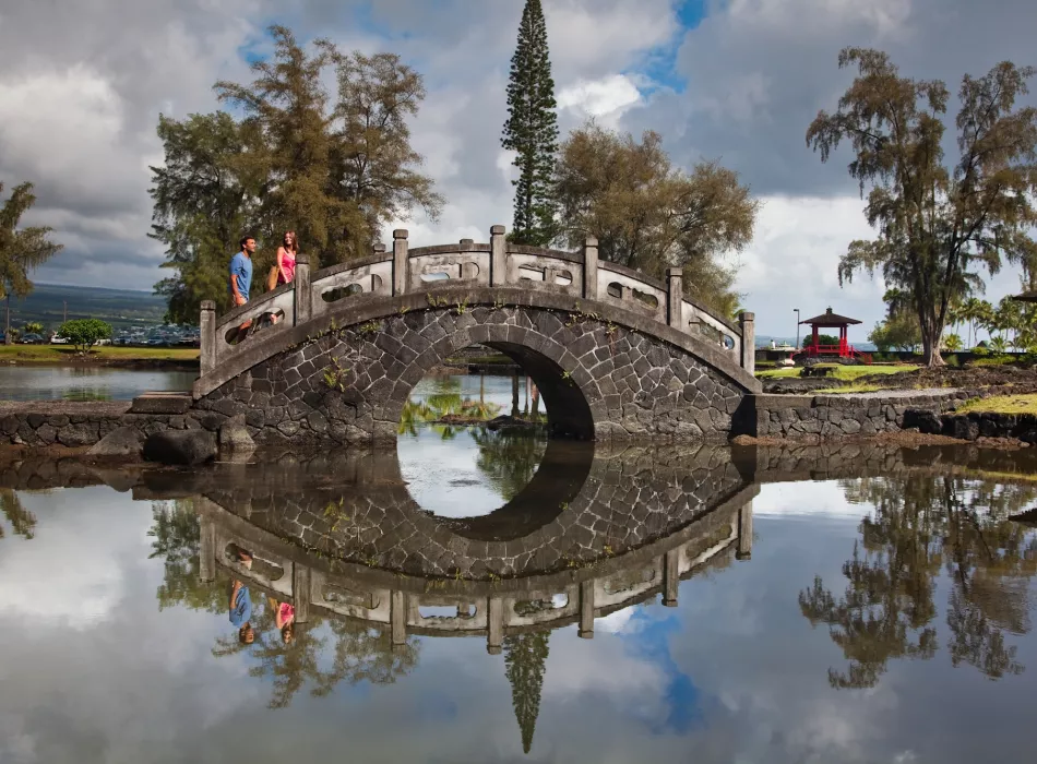 Stone arch bridge over a reflective pond in Liliuokalani Gardens with trees, a pagoda, and visitors in the background.