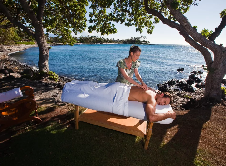 An outdoor massage setup under a tree by the ocean, with a therapist giving a massage on a sunny day.
