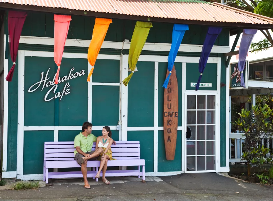 Couple sitting on a purple bench outside the Holualoa Cafe, a charming coffee shop adorned with colorful flags and a surfboard sign.