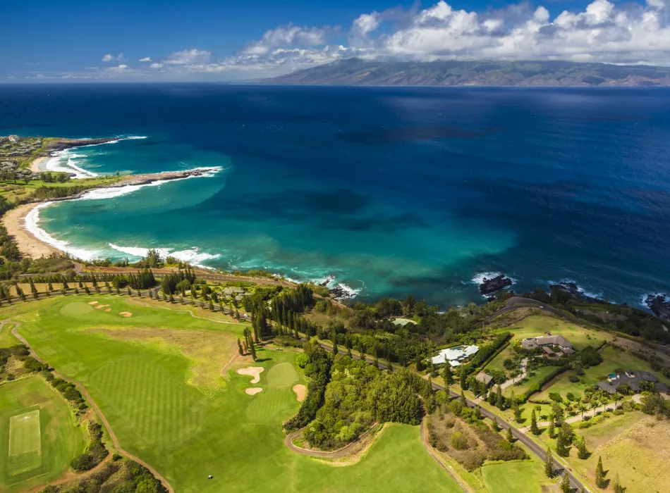 Aerial view of a scenic golf course with lush greenery along the coastline, overlooking turquoise waters and distant islands.