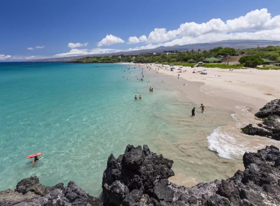 Visitors enjoying the turquoise waters and white sands of Hapuna Beach State Park with a rocky volcanic coastline in the foreground.