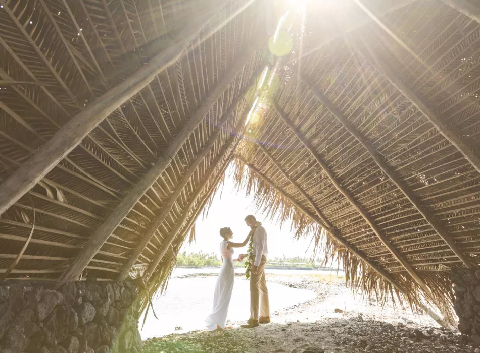 A couple stands under a traditional palm-frond structure by the water, bathed in sunlight.