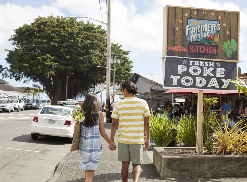 Couple walking toward a market with a sign reading 'Fresh Poke Today' and stalls selling local goods and food in the background.