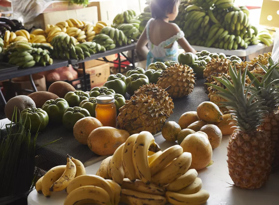 Table filled with tropical fruits such as bananas, pineapples, and papayas at the Hilo Farmers Market, with a child in the background.