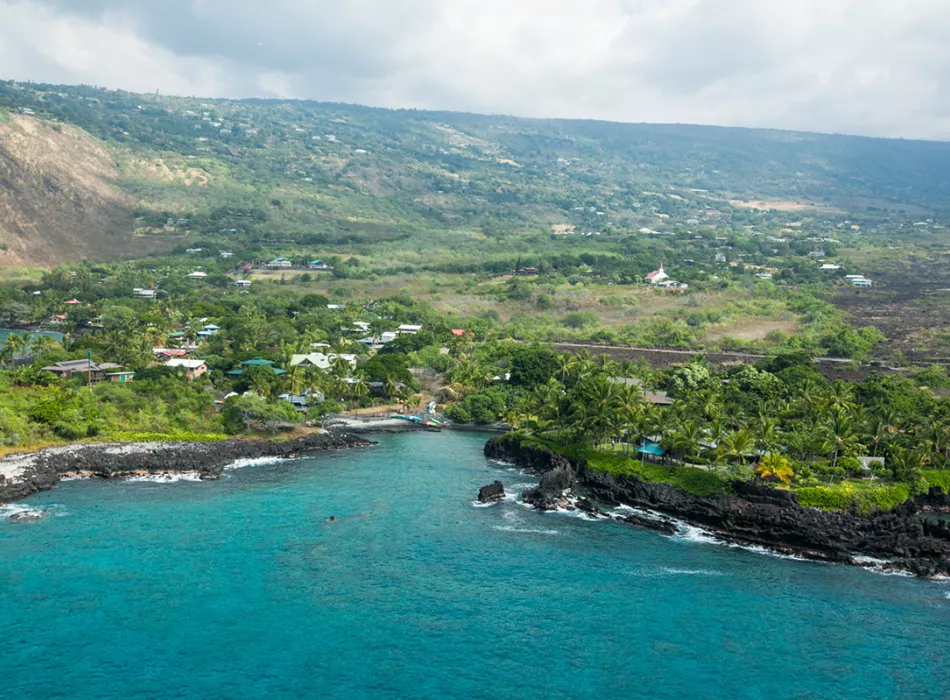 Aerial view of Kealakekua Bay with turquoise waters, lush greenery, rocky cliffs, and scattered buildings along the shoreline.