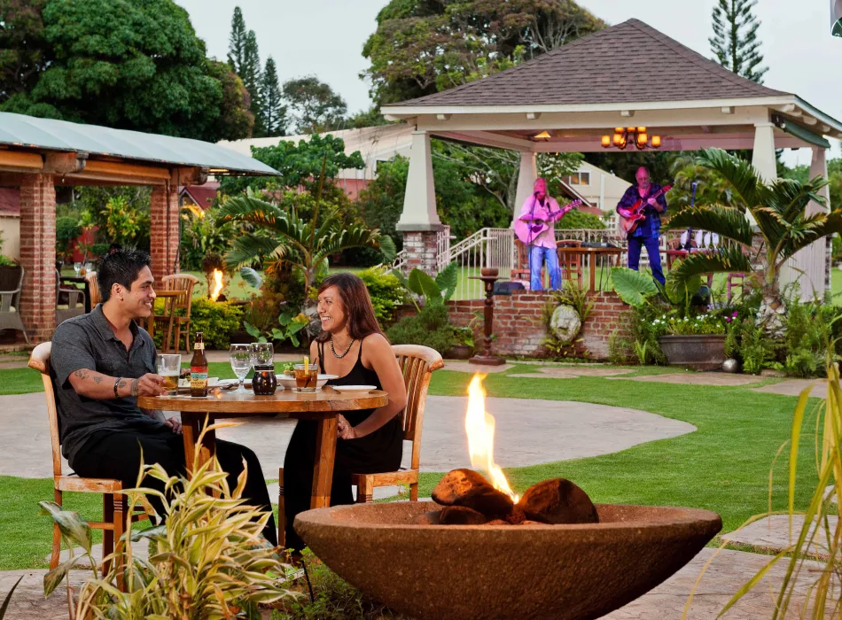 A couple dining outdoors near a firepit, with live musicians playing on a raised stage surrounded by greenery and tropical plants.