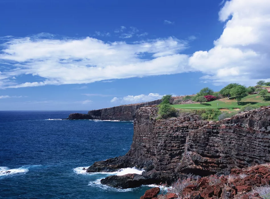 Cliffside golf course with lush greenery overlooking the ocean under a bright blue sky with scattered clouds.