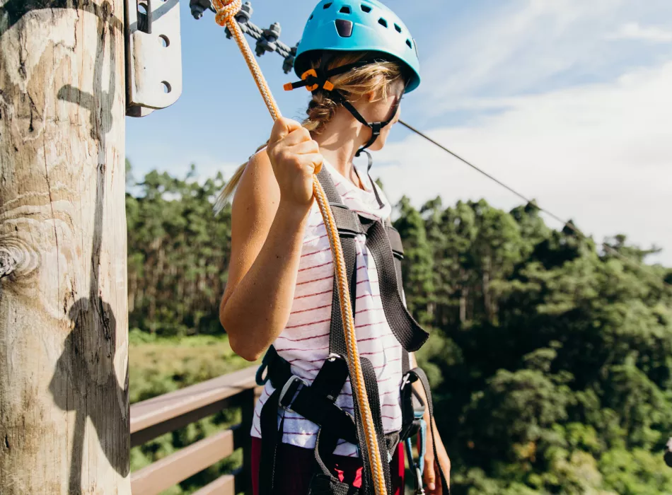 Woman wearing a blue helmet and harness holds a rope, overlooking a tropical landscape.