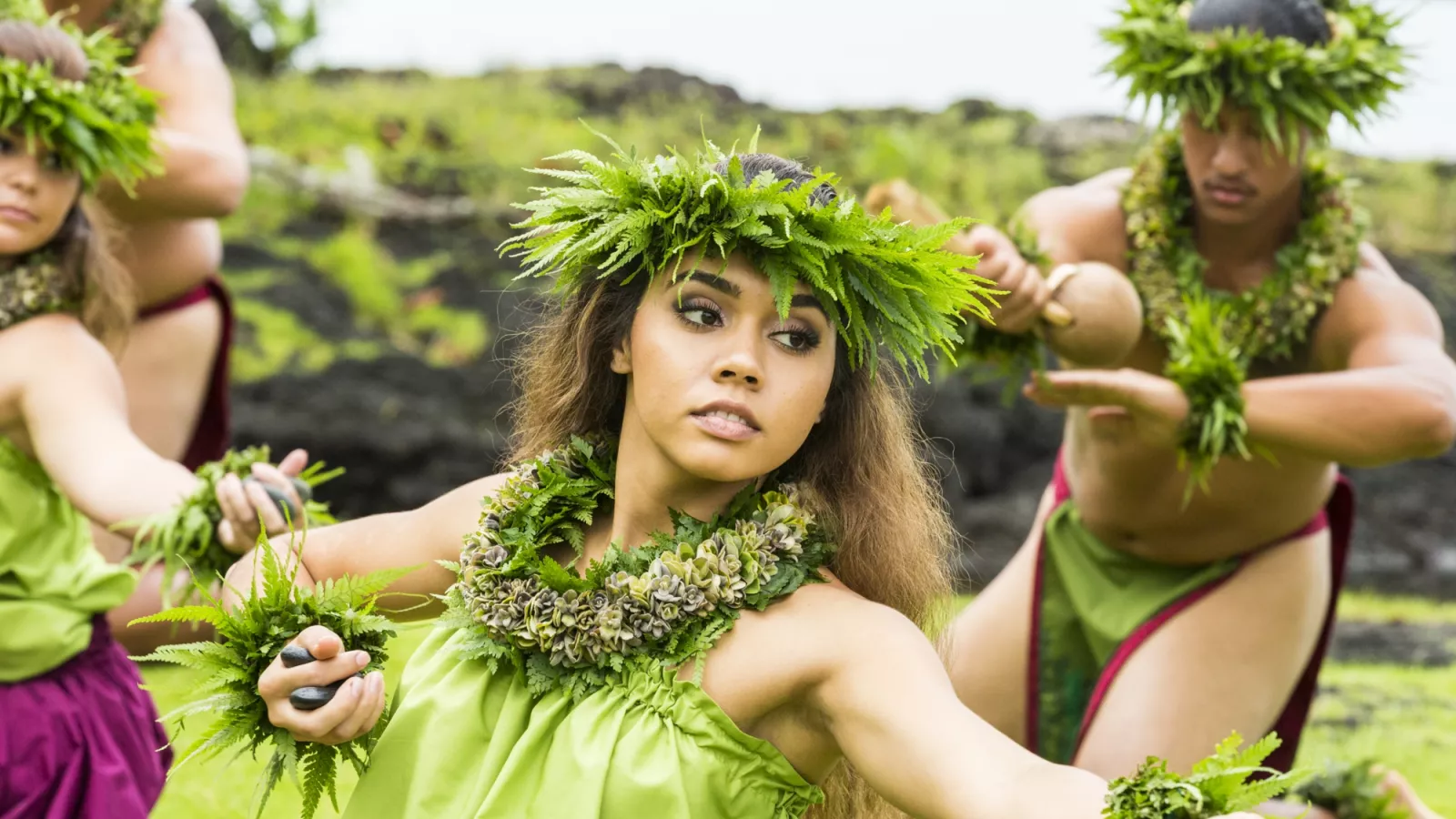 A Hawaiian dancer wearing a green dress and leaf crown performing hula, with another dancer holding a gourd instrument in the background.