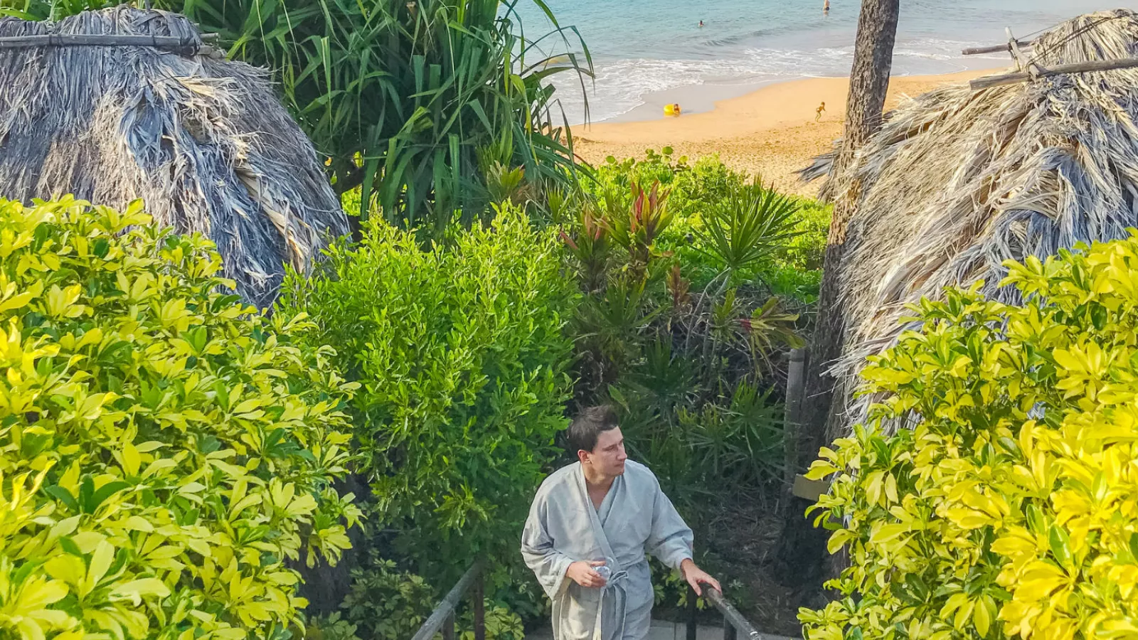 A man in a gray robe walks up stairs between thatched huts with a beach in the background.